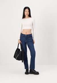 Witte lange mouwen crop top gecombineerd met donkerblauwe jeans en zwarte chunky laarzen. Model houdt een zwarte tote bag vast. Eenvoudig, strak ontwerp.