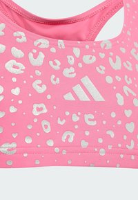 Rozā sporta krūšturis ar strukturētu leopardu rakstu sudraba mirdzumā, centrā ar Adidas logotipu un izgriezumu pie kakla.