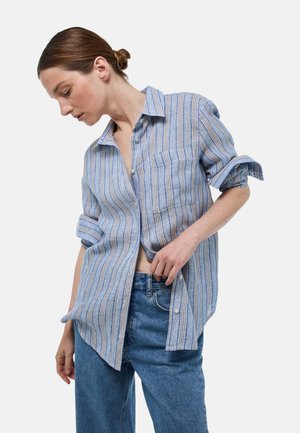 Chemise à boutons bleu et blanc rayée en tissu léger, avec une poche et des manches retroussées. Associée à un jean bleu.