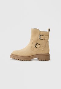 Bottines en daim beige avec deux sangles à boucle réglables, une semelle en caoutchouc brun épais et une texture lisse.