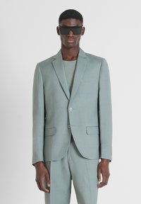 Completo in lino verde chiaro con giacca a un bottone, due bottoni e tasche a pattina. Il modello indossa occhiali da sole scuri, mostrando un look moderno.