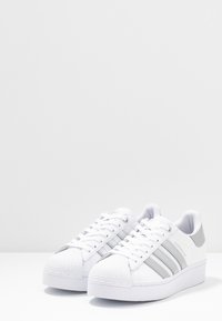 Bílé tenisky s texturovaným gumovým špičkou, hladkým koženým svrškem, stříbrným designem se třemi pruhy a polstrovaným límcem.