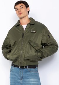 Olive green bomber jacket with a zip front, wide collar, and two large front pockets. Features a branded patch and ribbed cuffs.