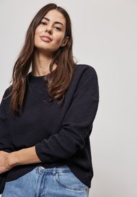 Navy Pullover mit Fischgrätenmuster, Rundhalsausschnitt und langen Ärmeln. Kombiniert mit hellblauen Jeans. Weiche Textur. Lässiger Schnitt.