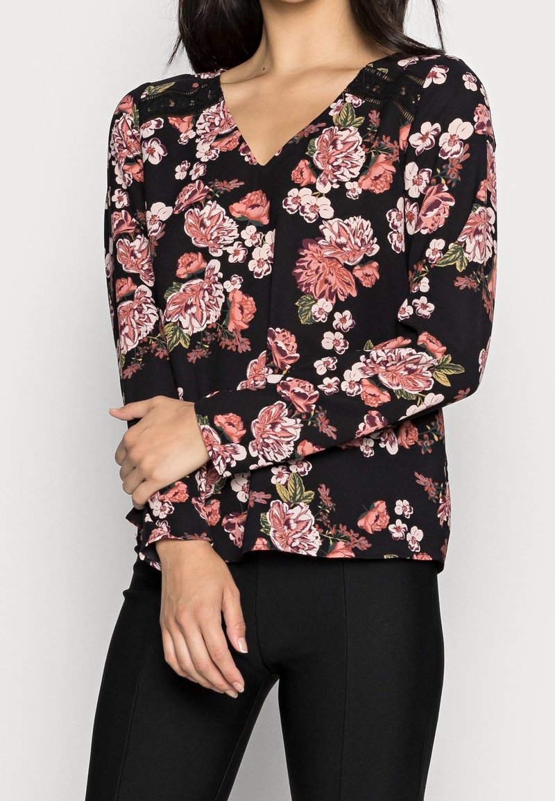 Femme portant un chemisier noir à manches longues avec des motifs floraux roses et blancs et un pantalon noir ajusté, les bras croisés à la taille.