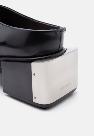 Μαύρο δερμάτινο παπούτσι με ψηλό τετράγωνο τακούνι σε μορφή μπλοκ που διαθέτει αντανακλαστική ασημί μεταλλική πλάκα στο πίσω μέρος με ανάγλυφο κείμενο.