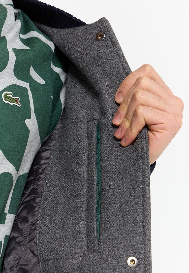 Veste grise en tissu lisse avec une poche zippée verte cachée à l'intérieur. Comprend une fermeture à pression et un col côtelé.