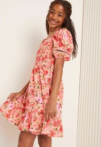 Blumenkleid mit gerafftem Oberteil, kurzen Puffärmelchen und einem rüschengesäumten Saum. Mit einem rosa Grundton und roten sowie rosa Blumenmustern.