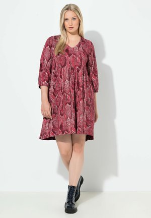 Rotes Kleid mit Schlangenmustern, V-Ausschnitt, Dreiviertelärmeln und einem schwingenden Rock. Das Modell trägt schwarze Schnürstiefel.