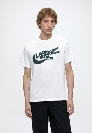 Junger Mann trägt ein weißes T-Shirt mit einer großen schwarzen Krokodil-Grafik und dem Wort „Lacoste“, kombiniert mit schwarzen Hosen.