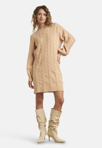 Abito maglione in morbido beige con motivo a trecce, maniche lunghe e vestibilità ampia, abbinato a stivali al ginocchio dall'aspetto morbido.