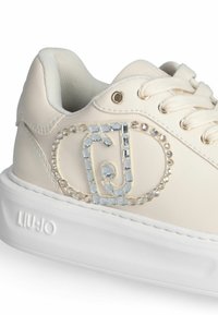 Baskets en cuir beige avec une semelle en caoutchouc blanc brillant. Présente un logo décoratif entouré de strass sur le côté et des lacets beiges.