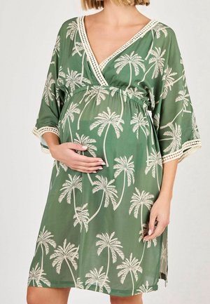 Femme enceinte dans une robe de maternité verte avec des motifs de palmier blancs, tenant son ventre, se tenant devant un fond uni.