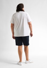 Witte katoenen t-shirt, korte mouwen, ontspannen pasvorm, gecombineerd met marineblauwe shorts. Model staat casual, met witte sneakers. Schoon achtergrond.