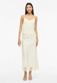 Kvinne iført kremhvit satengslipskjole med blonder, tynne stropper og sølvfargede åpen-tå hæler, stående foran en hvit bakgrunn.