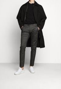 Cappotto oversize nero sopra una camicia nera aderente, pantaloni grigi a fantasia e sneakers bianche; texture di tessuto lisce e vestibilità su misura.