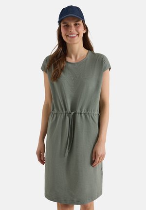 Smilende kvinde iført en afslappet grønstribet kjole med bindebånd i taljen og en mørkeblå baseballkasket, stående mod en ensfarvet baggrund.