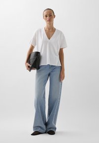 Witte T-shirt met korte mouwen en V-hals, gecombineerd met lichte blauwe wijde broek. Het model houdt een zwarte tas vast. Glad materiaal en ontspannen pasvorm.