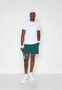 Valge sportlik t-särk logoga, rohelised lühikesed püksid, valged sokid ja tennisetossud. Hoidmas musta ja sinist tennisereketit. Lihtne, sportlik disain.