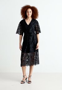 Robe en dentelle noire avec des manches courtes, décolleté en V, taille cintrée et un ourlet juste en dessous du genou. Élevée par des chaussures à talons avec des accents décoratifs.