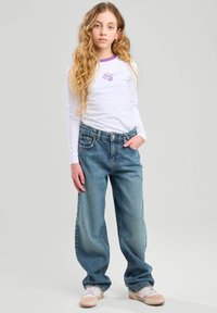 Camicia bianca a maniche lunghe con accenti viola e logo, abbinata a jeans larghi blu chiaro. Sneakers bianche con dettagli rosa.