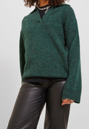 Femme portant un pull vert foncé en tissu pelucheux avec col, et un pantalon en cuir noir, accessoirisée d'un collier chaîne argenté épais.