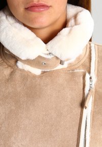 Beige faux suede jacket with a plush white fur collar, side zipper, and contrasting white stitching along the edges. Soft texture.