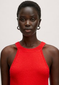 Red knitted sleeveless top with a ribbed neckline and textured fabric. Features a high neck and wide armholes.