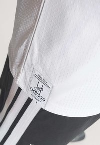 Gros plan sur un vêtement de sport Adidas blanc avec une étiquette indiquant un partenariat avec l'équipe nationale allemande depuis 1954, pantalon rayé noir et blanc visible.