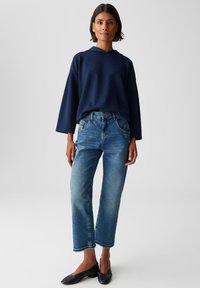 Donna in piedi con un abbigliamento casual, indossa un felpa blu navy larga, jeans blu a vita alta, e scarpe nere basse con tacco contro uno sfondo neutro.