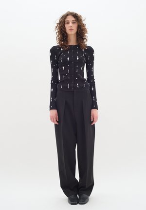 Schwarze gerippte Strickjacke mit silbernen Verzierungen, kombiniert mit hoch taillierten schwarzen Hosen. Das Model steht vor einem neutralen Hintergrund.