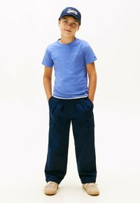 Ragazzo che indossa una maglietta blu Tommy Hilfiger, pantaloni cargo navy, sneakers beige e un cappellino navy Tommy, in piedi con le mani nelle tasche.