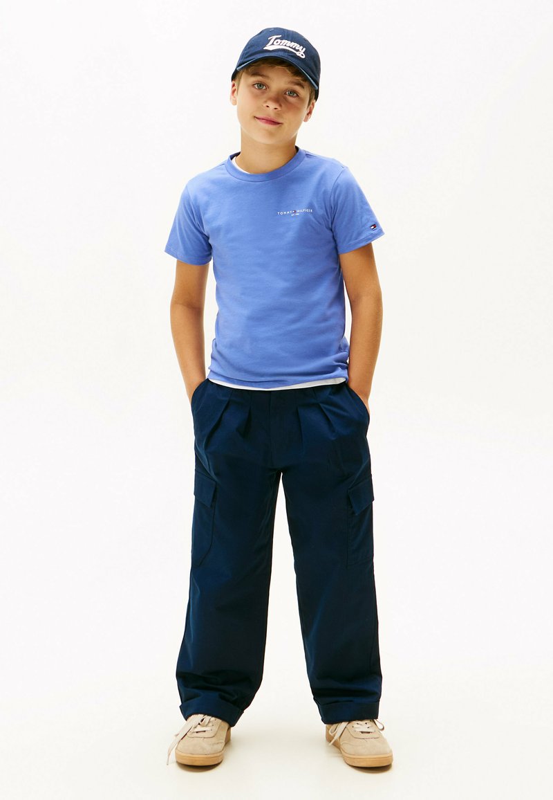 Ragazzo che indossa una maglietta blu Tommy Hilfiger, pantaloni cargo navy, sneakers beige e un cappellino navy Tommy, in piedi con le mani nelle tasche.