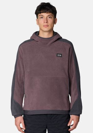 Junger Mann, der einen zweifarbigen lila Fleece-Hoodie mit hohem Kragen und dem Mountain Hardwear-Logo auf der Brust trägt, vor einem einfarbigen Hintergrund stehend.