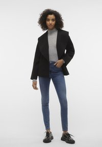 Svart skräddarsydd jacka med breda slag, lager över en grå polotröja, kombinerad med blå slim-fit jeans och svarta platåskor.