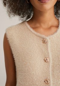 Top in maglia senza maniche beige con una texture morbida e pelosa, dotato di tre bottoni dorati decorativi a torsione e un collo rotondo.