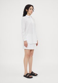 Camicia bianca di lino con chiusura a bottoni e maniche lunghe, abbinata a una gonna bianca e sandali neri a due fasce, su uno sfondo semplice.