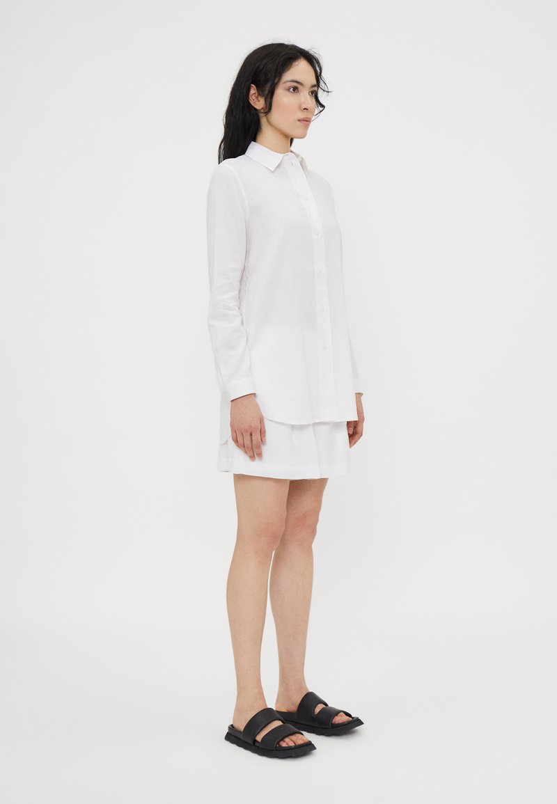 Camicia bianca di lino con chiusura a bottoni e maniche lunghe, abbinata a una gonna bianca e sandali neri a due fasce, su uno sfondo semplice.