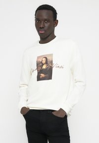 Cremefarbener Sweatshirt mit einem zentralen Druck der Mona Lisa und dem Text "Leonardo Da Vinci" darunter. Kombiniert mit schwarzen Jeans.