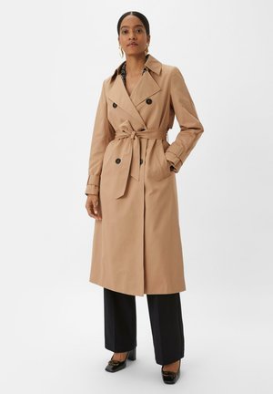 Beige trench coat with double-breasted design, belted waist, and large buttons. Model wears black wide-leg trousers and heels.