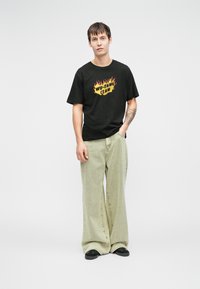 Μαύρη βαμβακερή μπλούζα T-shirt με το "WU-TANG CLAN" σε πορτοκαλί φλόγες, συνδυασμένη με άνετο ανοιχτό πράσινο τζιν παντελόνι και μαύρα παπούτσια.