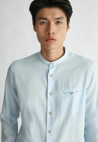 Camicia azzurro chiaro con colletto alla mandarina, dotata di taschino sul petto, tessuto testurizzato e bottoni in legno per la chiusura.