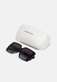 Ochelari de soare pătrați negri cu lentile întunecate cu gradient, așezați lângă o carcasă albă cu un logo negru "Calvin Klein Jeans".