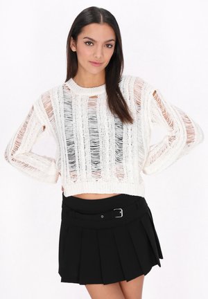 Maglione bianco tagliato con modelli di lavorazione a maglia orizzontali e verticali, caratterizzato da dettagli strappati. Abbinato a una gonna nera plissè e una cintura.