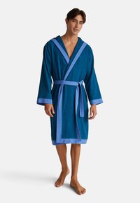 Jeune homme debout pieds nus portant un peignoir en teal avec une bordure bleu clair et une ceinture, contre un fond blanc uni.