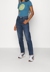 Blue short-sleeve t-shirt with a yellow floral design, paired with high-waisted dark blue jeans and white athletic shoes with black accents.