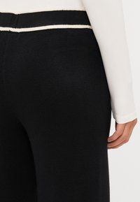 Černé pletené kalhoty s hladkou texturou, s kontrastním bílým horizontálním pruhem v pase. Boční pohled ukazuje splývání látky.