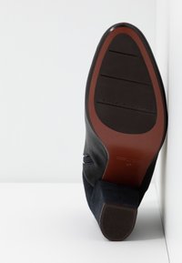 Semelle d'une bottine en cuir noir et suède avec talon bloc, montrant la bande de roulement marron et noire ainsi que le nom de la marque "Chie Mihara" embossé sur la semelle.