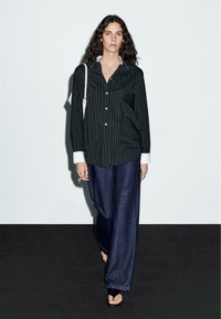 Donna che cammina con una camicia nera a righe verticali con polsini bianchi, indossando ampi jeans blu scuro, mentre porta una borsa bianca e cammina scalza su un pavimento nero.