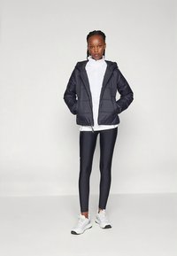 Černá prošívaná bunda s kapucí, pod ní bílý top s vysokým límcem a černé legíny. Modelka má na sobě bílé tenisky a stojí na jednobarevném pozadí.